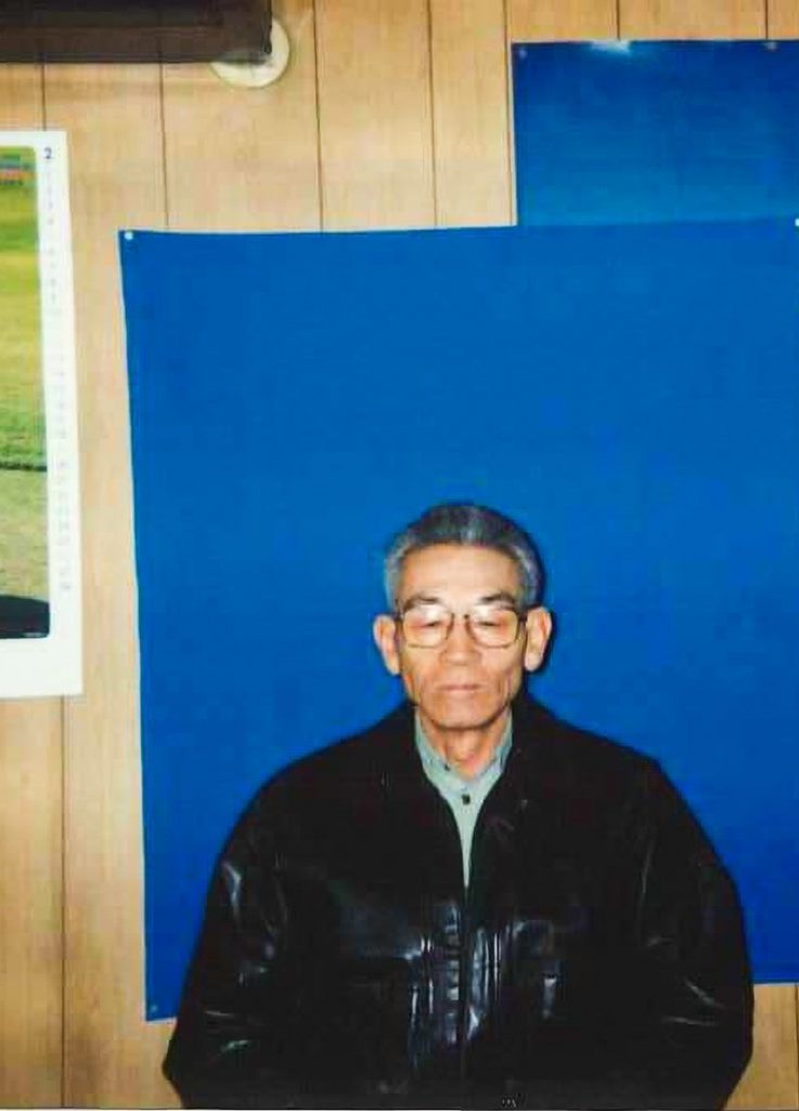事務所の壁を背に立つ、革のジャンパー姿の男性