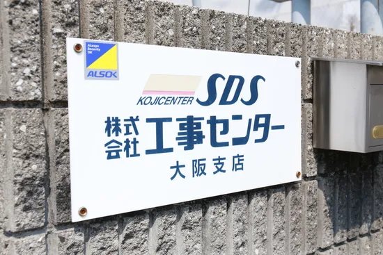 「株式会社工事センター 大阪支店」と書かれた社名看板が、ブロック塀に取り付けられている