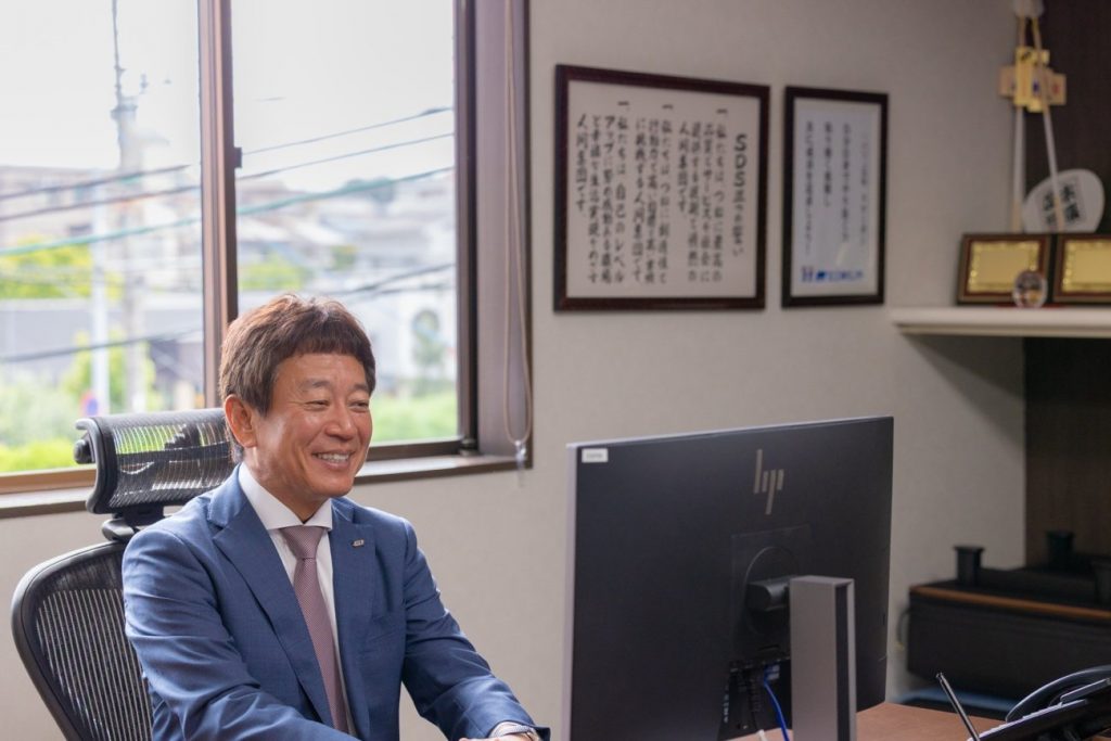 執務室のデスクでモニターに向かい、笑顔で話す末廣󠄁社長。壁に社訓の額が並ぶ